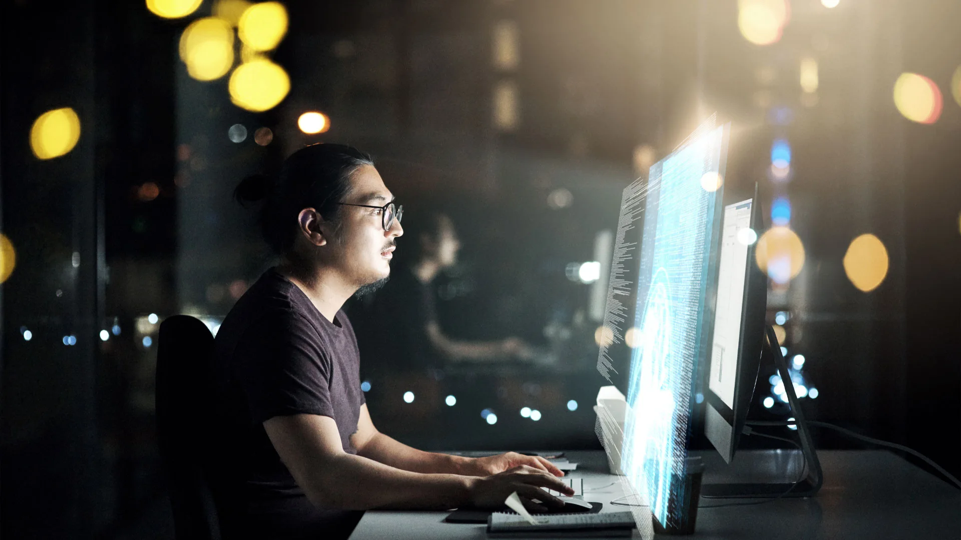 Software engineer, wearing glasses and surrounded by bokeh, looking at code on computer screen in a dark room.